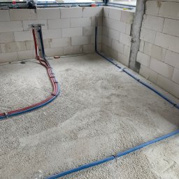 Marek Strzemiński - Instalacja rur CO i wodnych w surowym wnętrzu w Jeleniej Górze. Czerwone i niebieskie rury prowadzone po podłodze, przymocowane do ściany z białych bloczków.