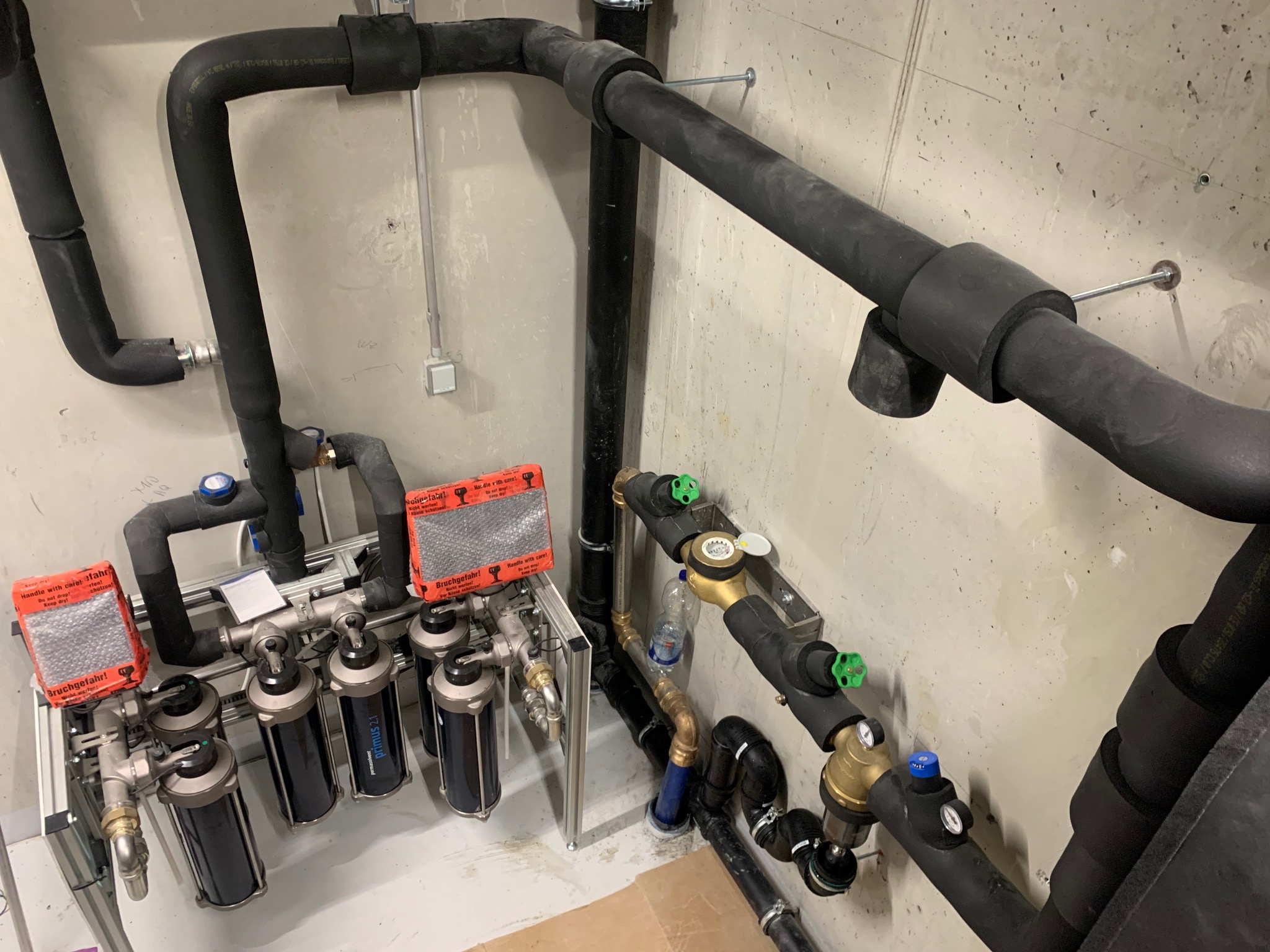 Złożona instalacja hydrauliczna z filtrami, rurami izolowanymi termicznie i zaworami w surowym wnętrzu. Widoczne elementy montażowe i zabezpieczenia.