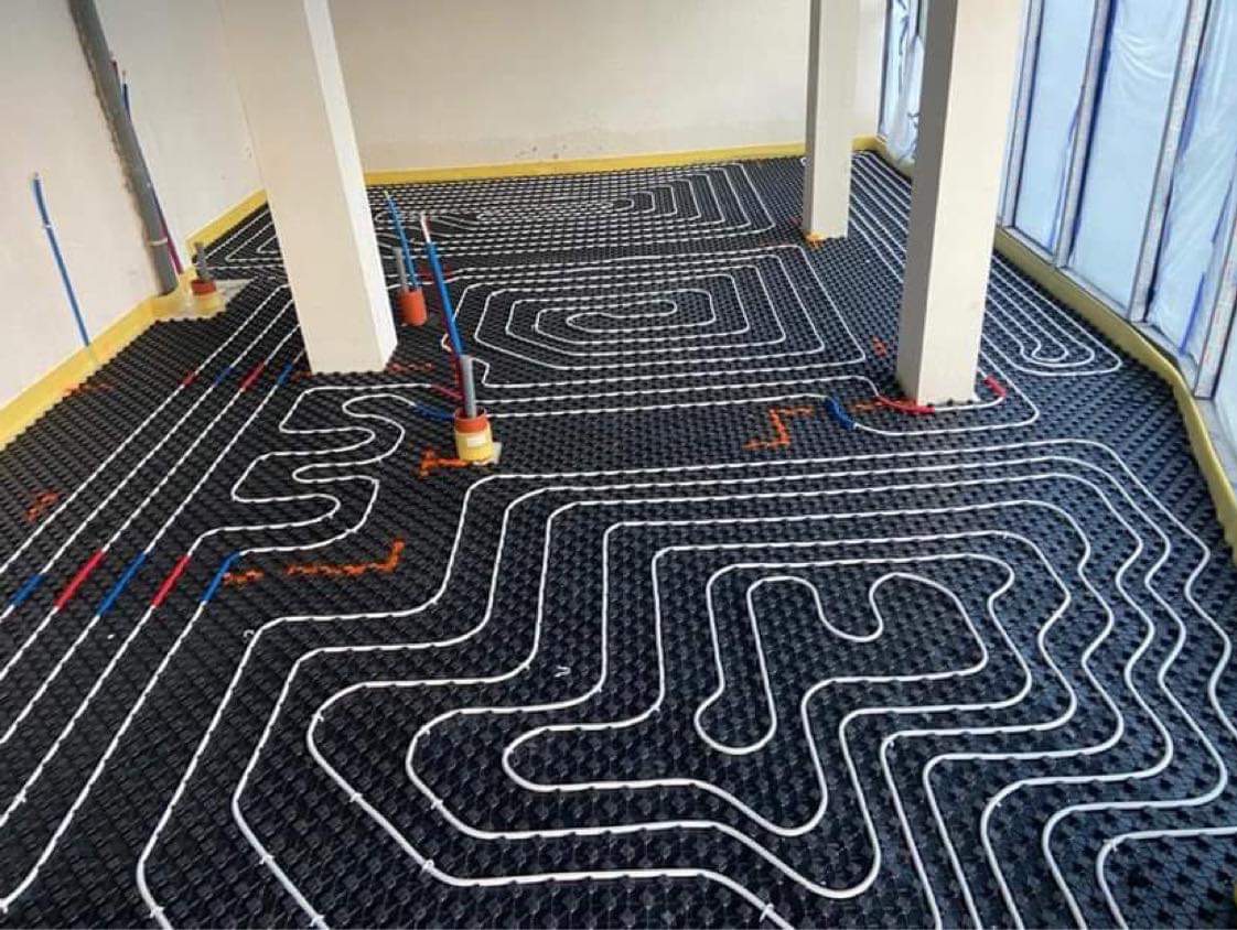 Białe rury ogrzewania podłogowego ułożone na czarnej macie izolacyjnej w przestronnym pomieszczeniu z kolumnami i oknami, przygotowane do zalania wylewką.