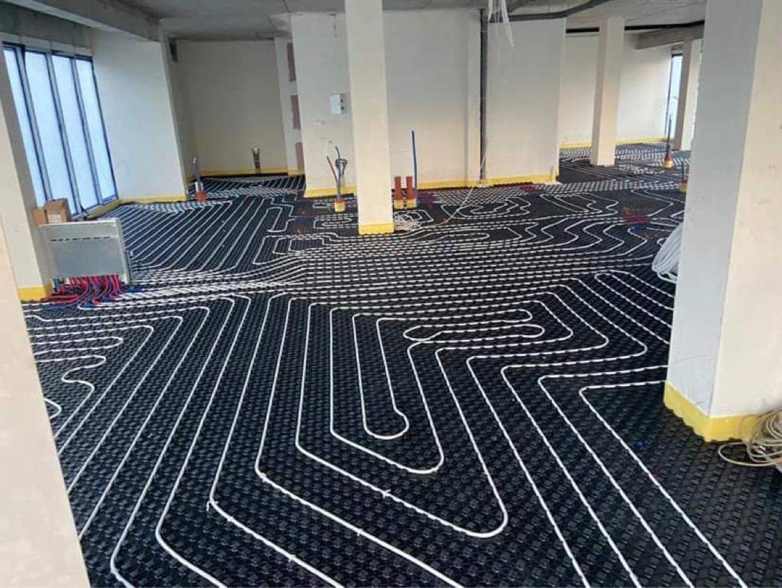 Rozłożona instalacja ogrzewania podłogowego z białych rurek na czarnej macie w surowym wnętrzu z kolumnami, gotowa do zalania. Widoczne okna i wystające elementy.