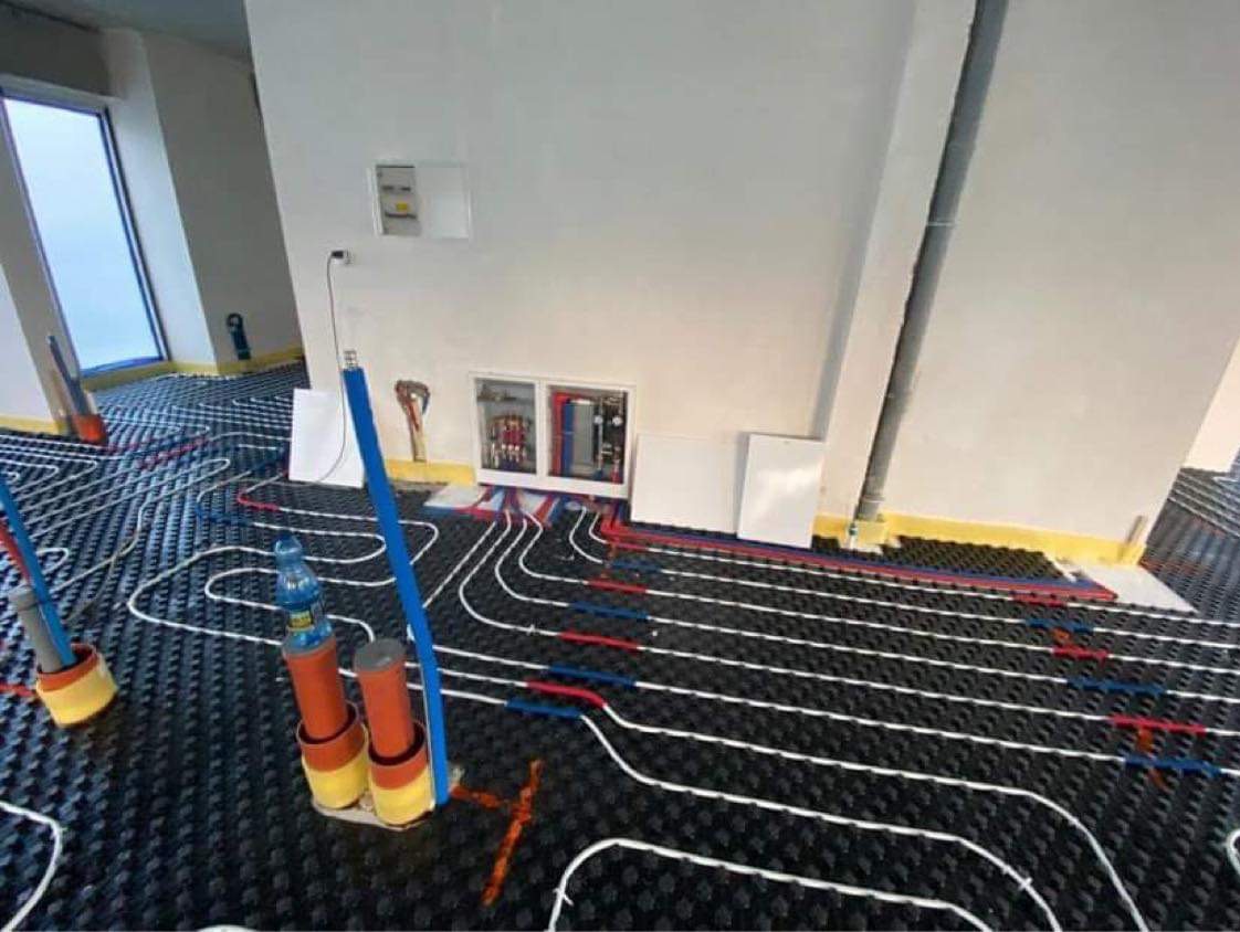 Instalacja ogrzewania podłogowego: rozłożone rury grzewcze (białe, czerwone, niebieskie) na czarnej macie izolacyjnej, widoczne elementy instalacji przy ścianie.