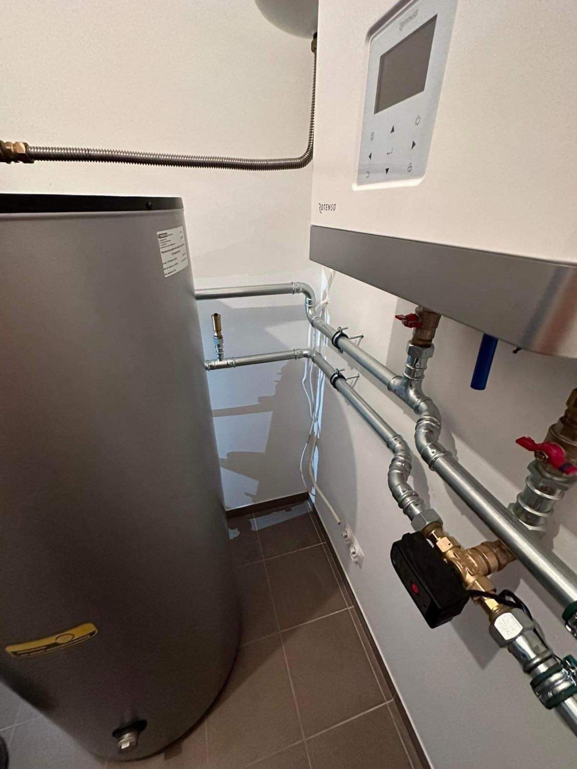 Szary zasobnik ciepłej wody użytkowej i biały piec gazowy Rotenso z podłączeniami hydraulicznymi na tle beżowej ściany i brązowych płytek podłogowych.