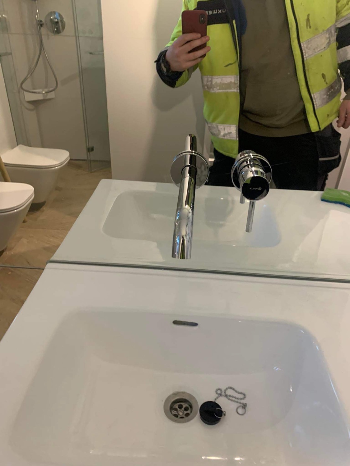 Nowa, chromowana bateria umywalkowa marki Kludi zamontowana w białej umywalce. Widoczne odbicie hydraulika w kamizelce podczas robienia zdjęcia telefonem.