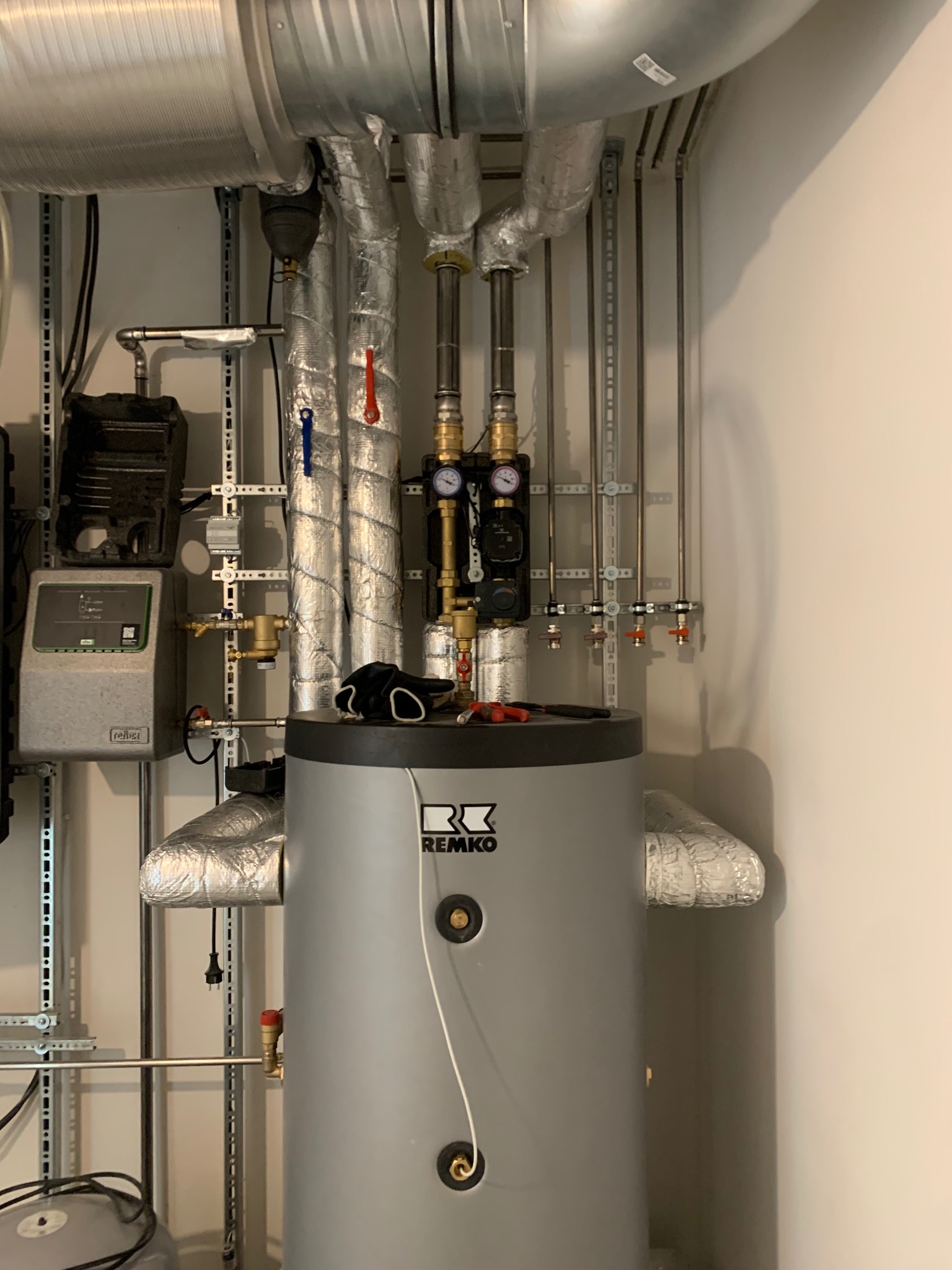 Szary zasobnik ciepłej wody REMKO z widocznymi instalacjami, rurami w izolacji i manometrami. Na zasobniku leżą rękawice robocze i narzędzia.