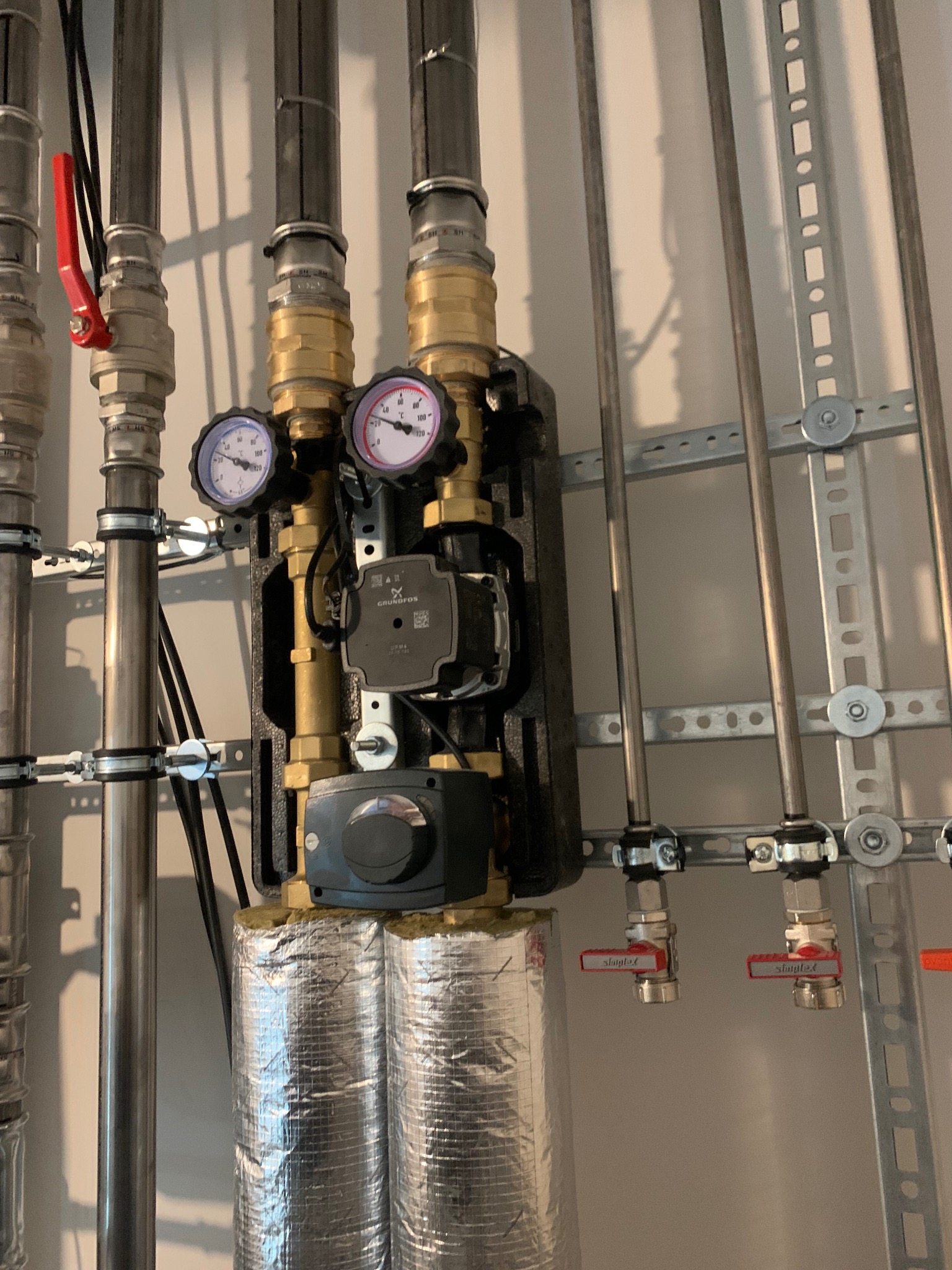 Węzeł cieplny z dwoma manometrami, pompą obiegową Grundfos i srebrną izolacją termiczną rur. Instalacja przymocowana do ściany za pomocą metalowych uchwytów.