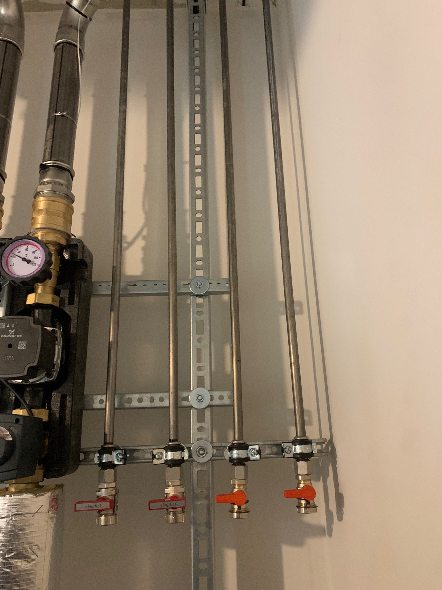 Zamontowane rury instalacji grzewczej z zaworami, przymocowane do ściany za pomocą metalowej listwy montażowej. Widoczny manometr i pompa obiegowa Grundfos.