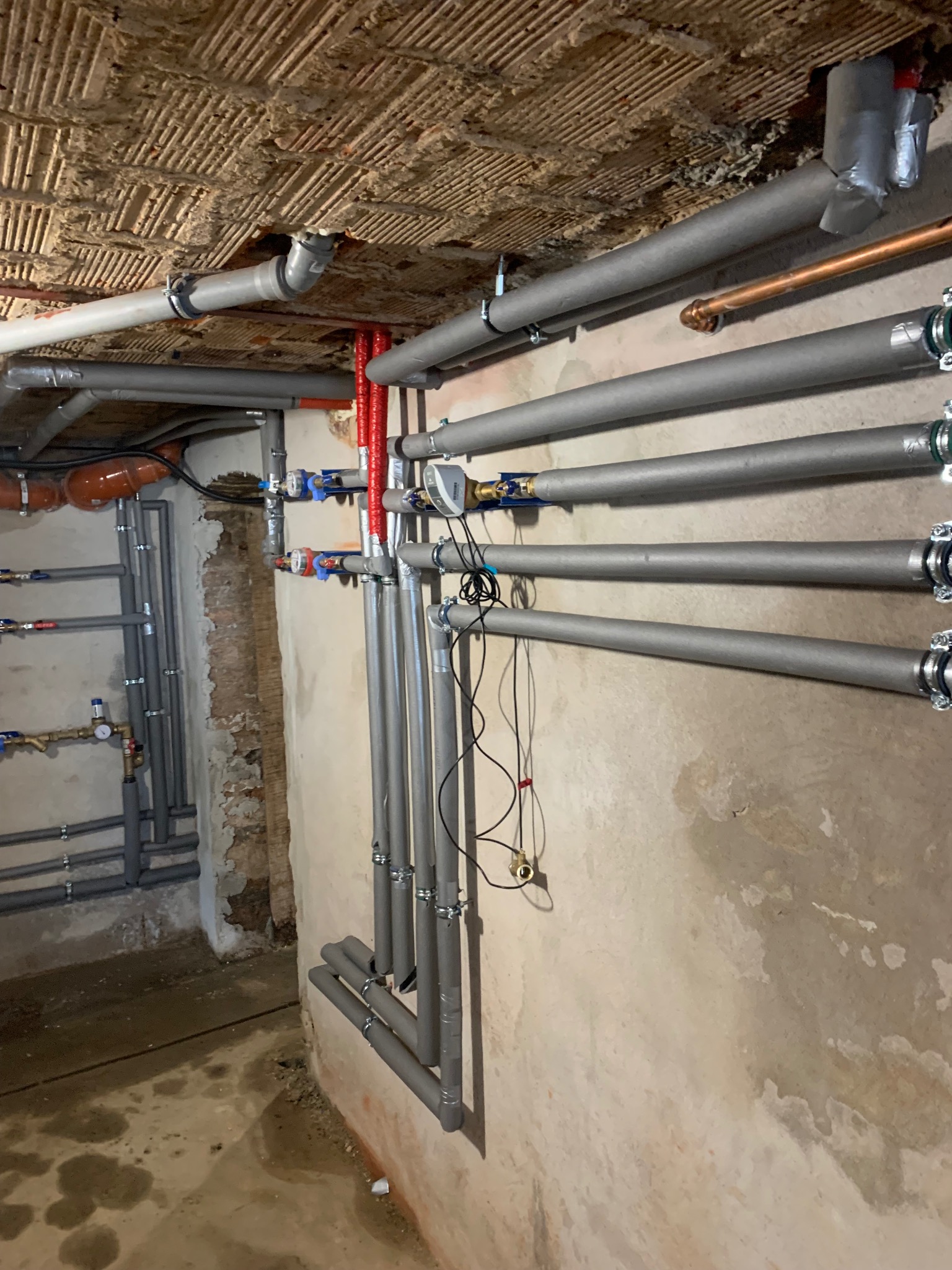 Szare, zaizolowane rury instalacji w surowym pomieszczeniu z widoczną konstrukcją stropu. Część rur z zaworami. Widoczne łączenia i mocowania.