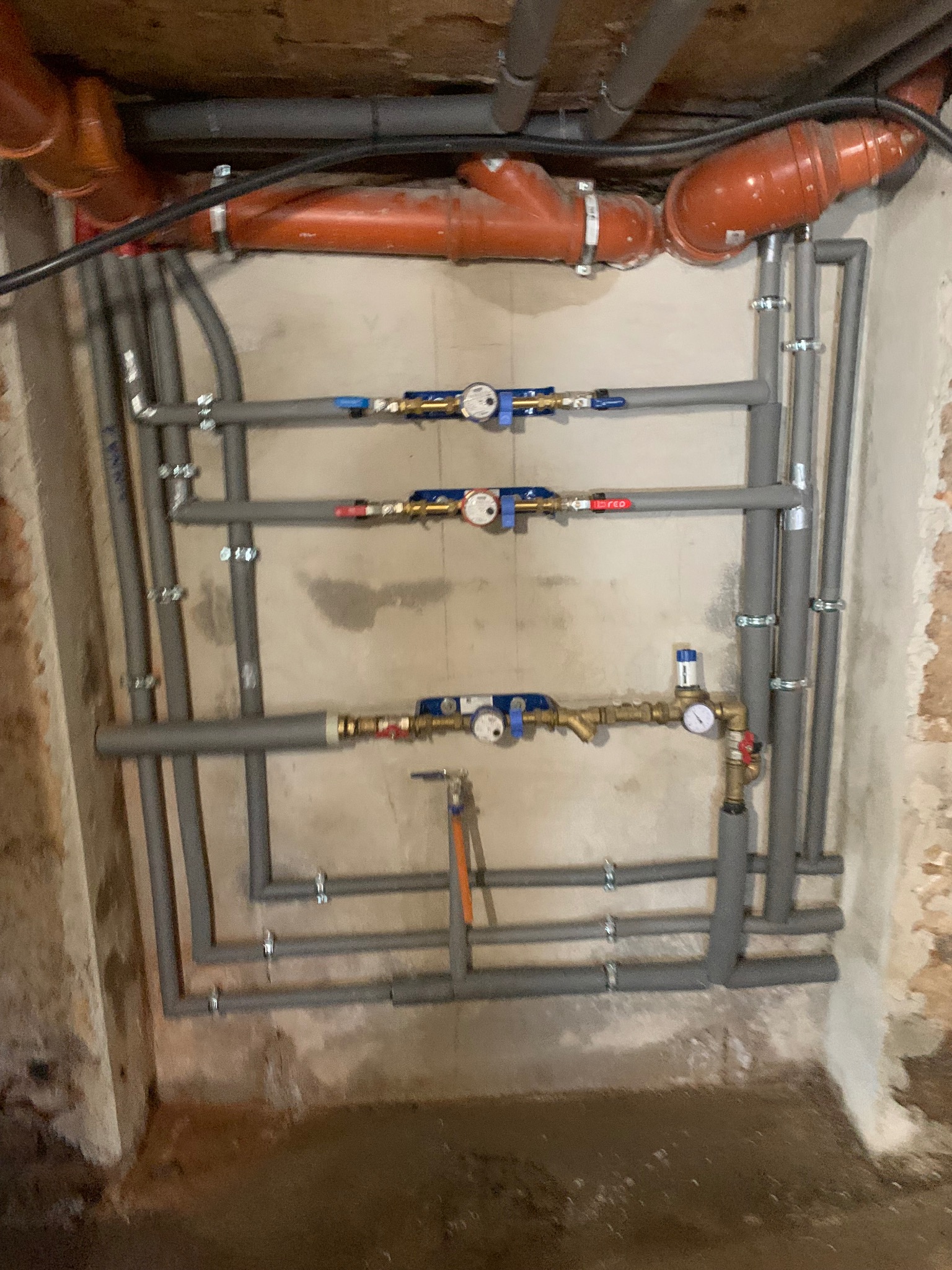 Szare rury instalacji hydraulicznej z zaworami i manometrami, zamocowane do ściany z widocznymi śladami wilgoci. U góry rury kanalizacyjne z pomarańczowego tworzywa.