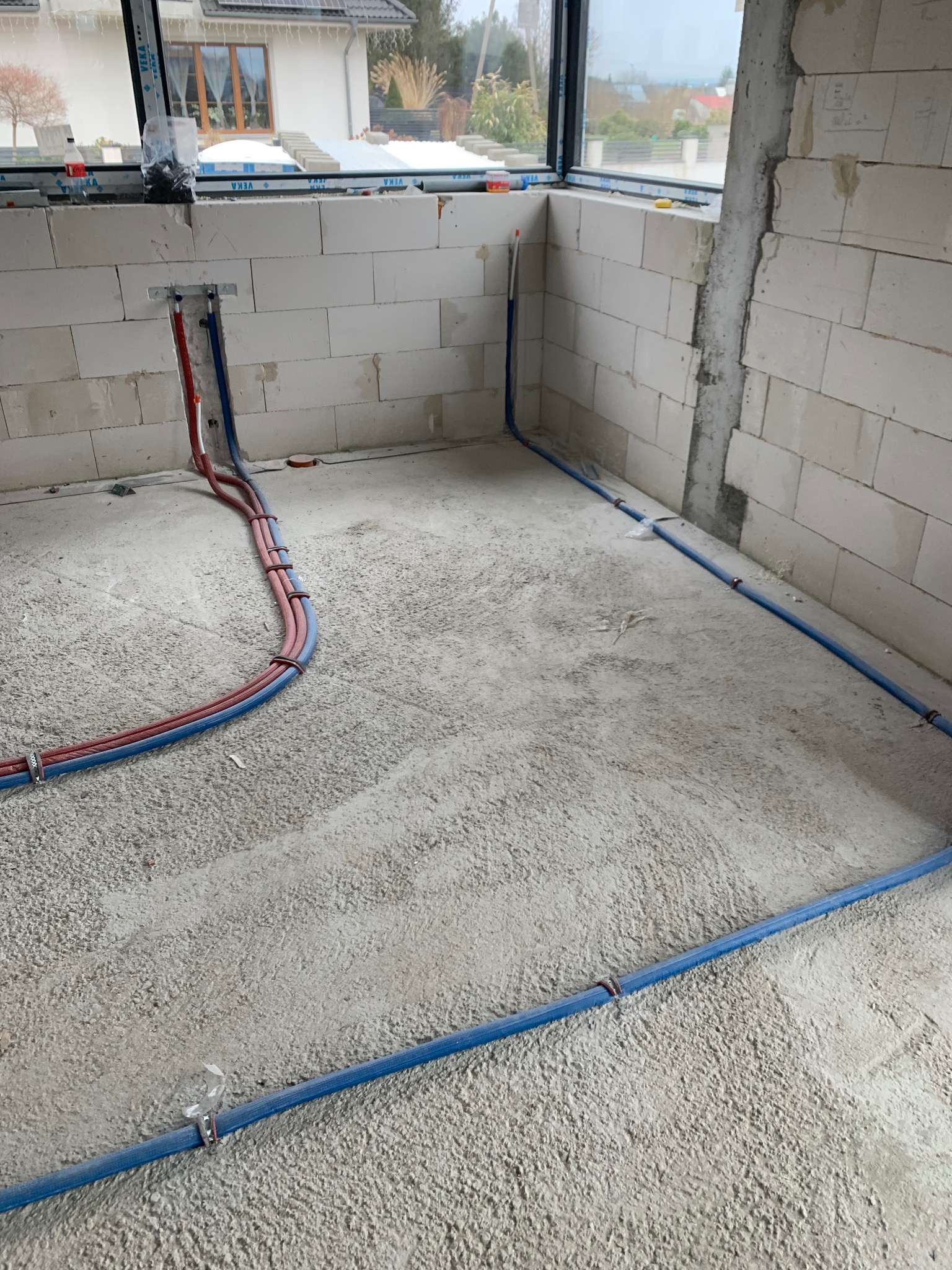 Instalacja rur CO i wodnych w surowym wnętrzu w Jeleniej Górze. Czerwone i niebieskie rury prowadzone po podłodze, przymocowane do ściany z białych bloczków.