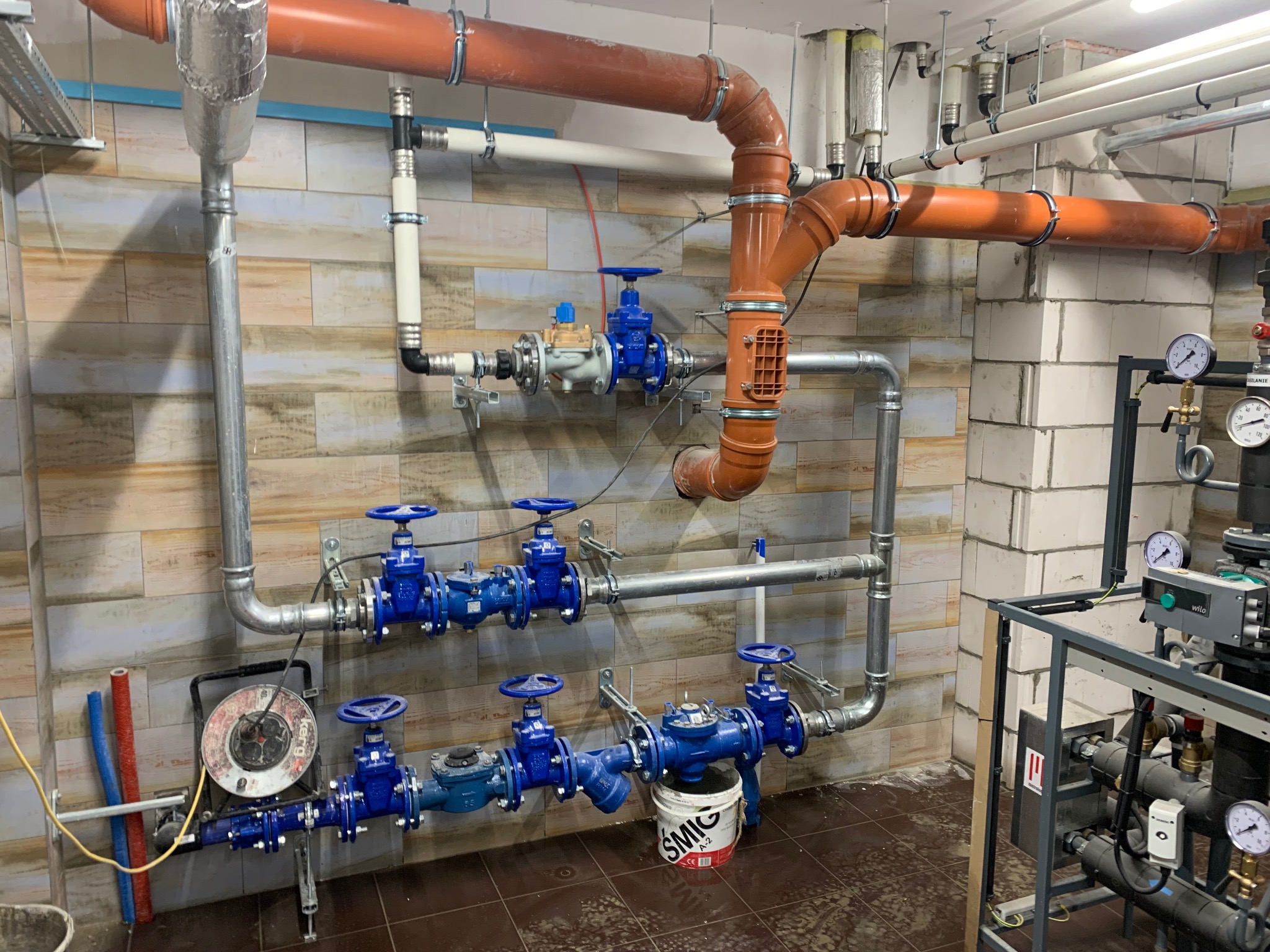 Kompleksowa instalacja hydrauliczna z rurami w kolorze pomarańczowym i srebrnym, zaworami z niebieskimi pokrętłami i manometrami, zamontowana na ścianie z płytek.