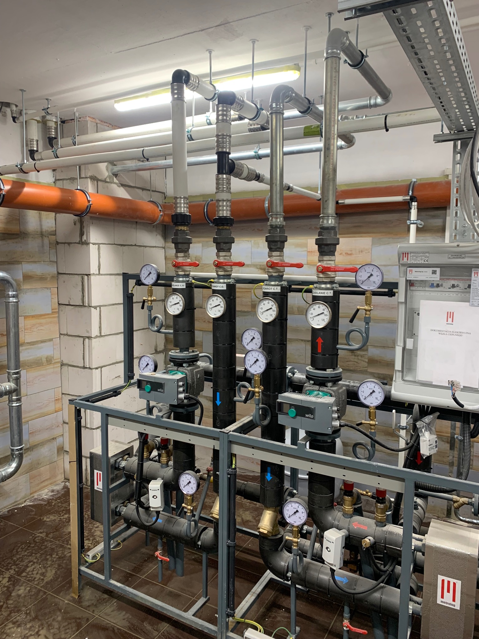 Kompleksowa instalacja grzewcza z rurami, zaworami i manometrami w kotłowni; widoczne elementy marki Wilo i Ferro. Instalacja w szarej ramie, na tle białej ściany z cegieł.