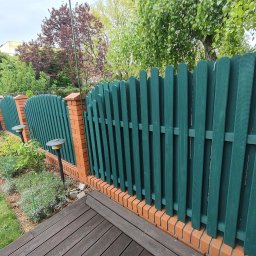 Renesans drewna - Zielony, drewniany płot sztachetowy z ceglaną podmurówką, oświetlony lampami ogrodowymi, oddzielający rabatę kwiatową od drewnianego tarasu.