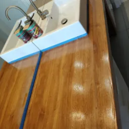 Błyszczący, świeżo polakierowany blat drewniany umywalki z dwiema puszkami lakieru i pędzlami. Zabezpieczony niebieską taśmą malarską. Widok z góry.