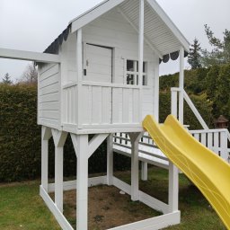 Renesans drewna - Biały drewniany domek zabaw dla dzieci z żółtą zjeżdżalnią, umieszczony w ogrodzie na trawie. Widoczne elementy konstrukcyjne i wykończeniowe.