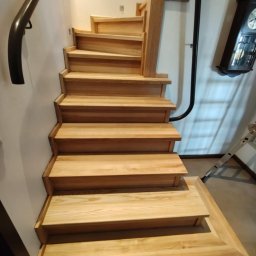 Renesans drewna - Jasne, drewniane schody z czarną, metalową poręczą, prowadzące w górę. Widoczny fragment ściany i zegar na ścianie. Solidne wykonanie, naturalne wykończenie drewna.