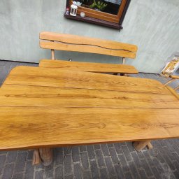 Renesans drewna - Drewniany stół i ławka ogrodowa z surowego drewna, ustawione na brukowanym tarasie. Widoczne detale usłojenia drewna i naturalne wykończenie. Na ścianie okno z dekoracjami.