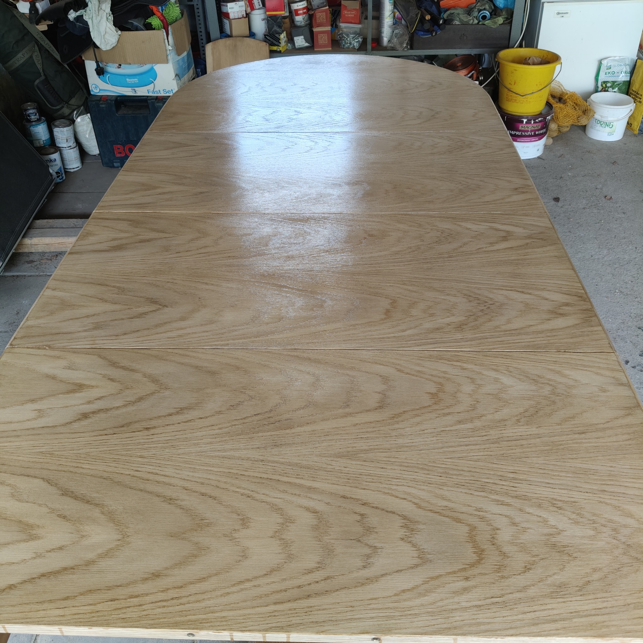 Odnowiony, rozkładany blat drewnianego stołu z widocznym rysunkiem słojów, po renowacji. Jasne drewno, gładka powierzchnia. W tle widoczne narzędzia i pojemniki.