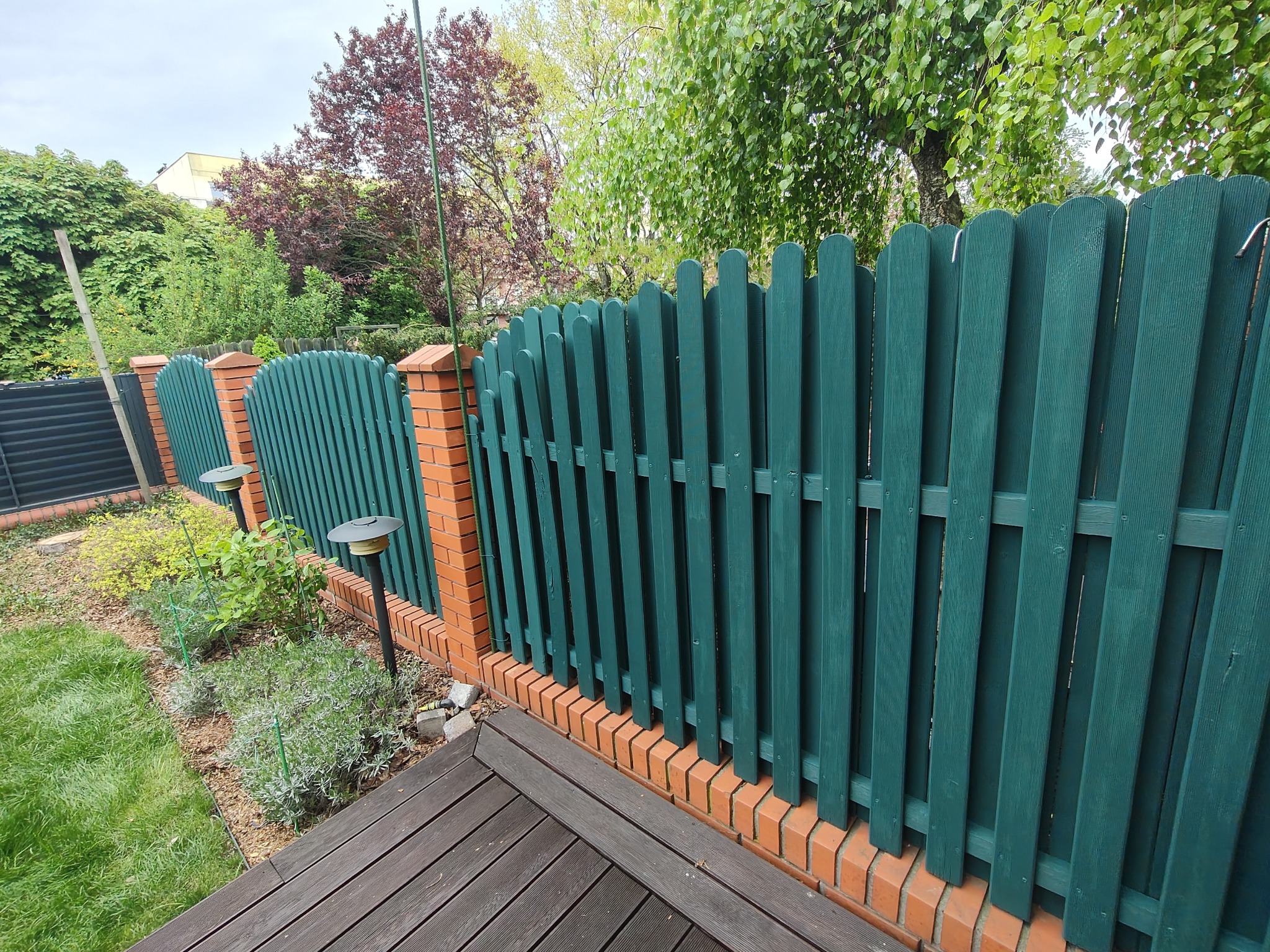 Zielony, drewniany płot sztachetowy z ceglaną podmurówką, oświetlony lampami ogrodowymi, oddzielający rabatę kwiatową od drewnianego tarasu.