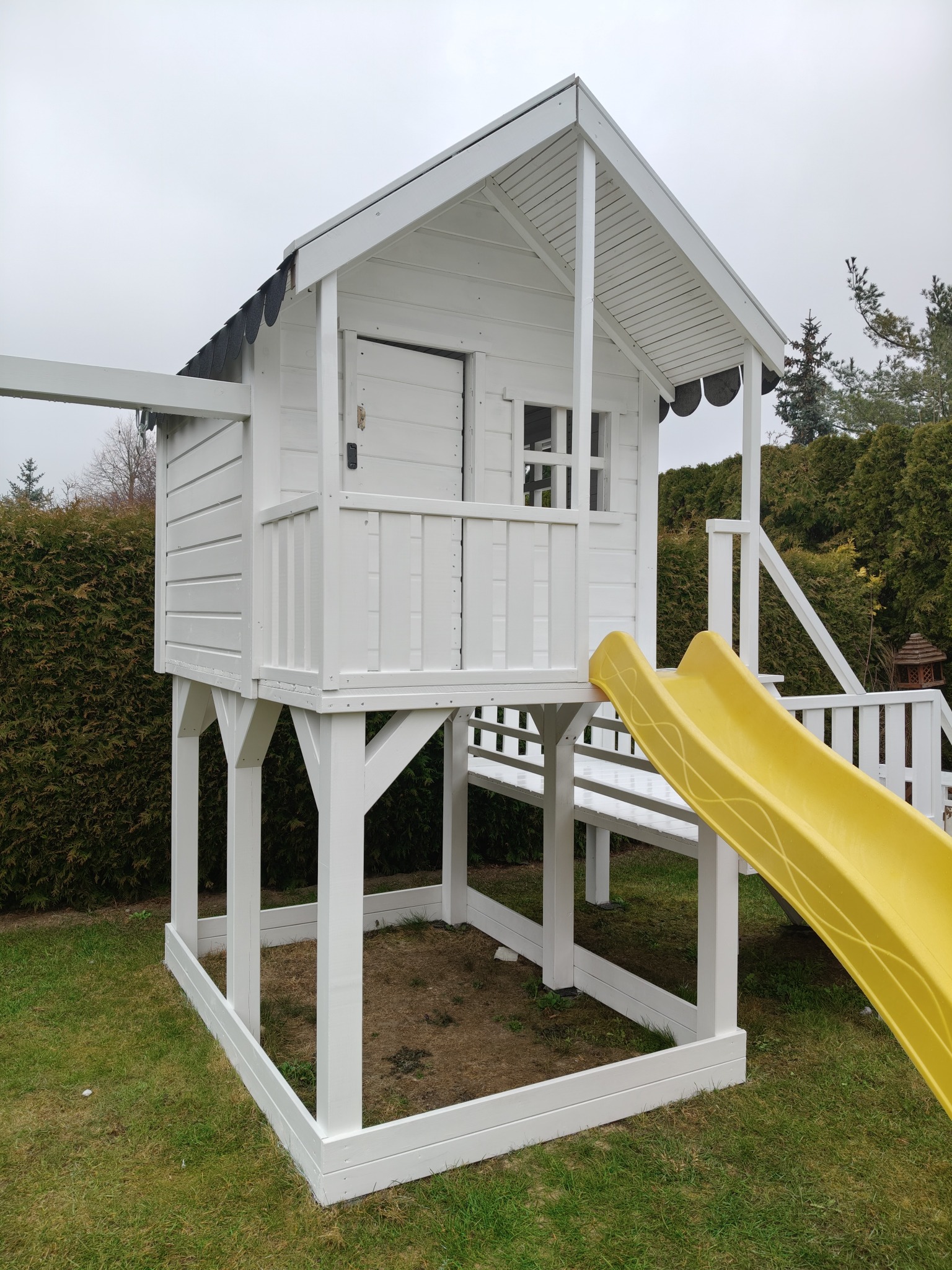 Biały drewniany domek zabaw dla dzieci z żółtą zjeżdżalnią, umieszczony w ogrodzie na trawie. Widoczne elementy konstrukcyjne i wykończeniowe.