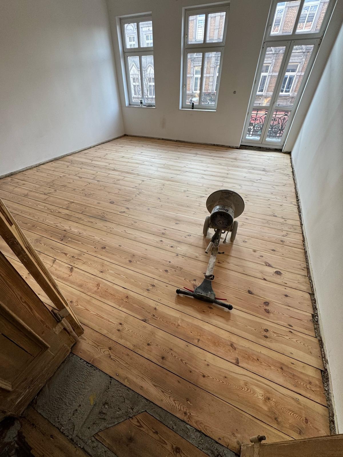 Cyklinowanie drewnianej podłogi w jasnym pomieszczeniu z oknami. Widoczna cykliniarka na tle odnowionej podłogi w trakcie prac. Fragment schodów z boku kadru.