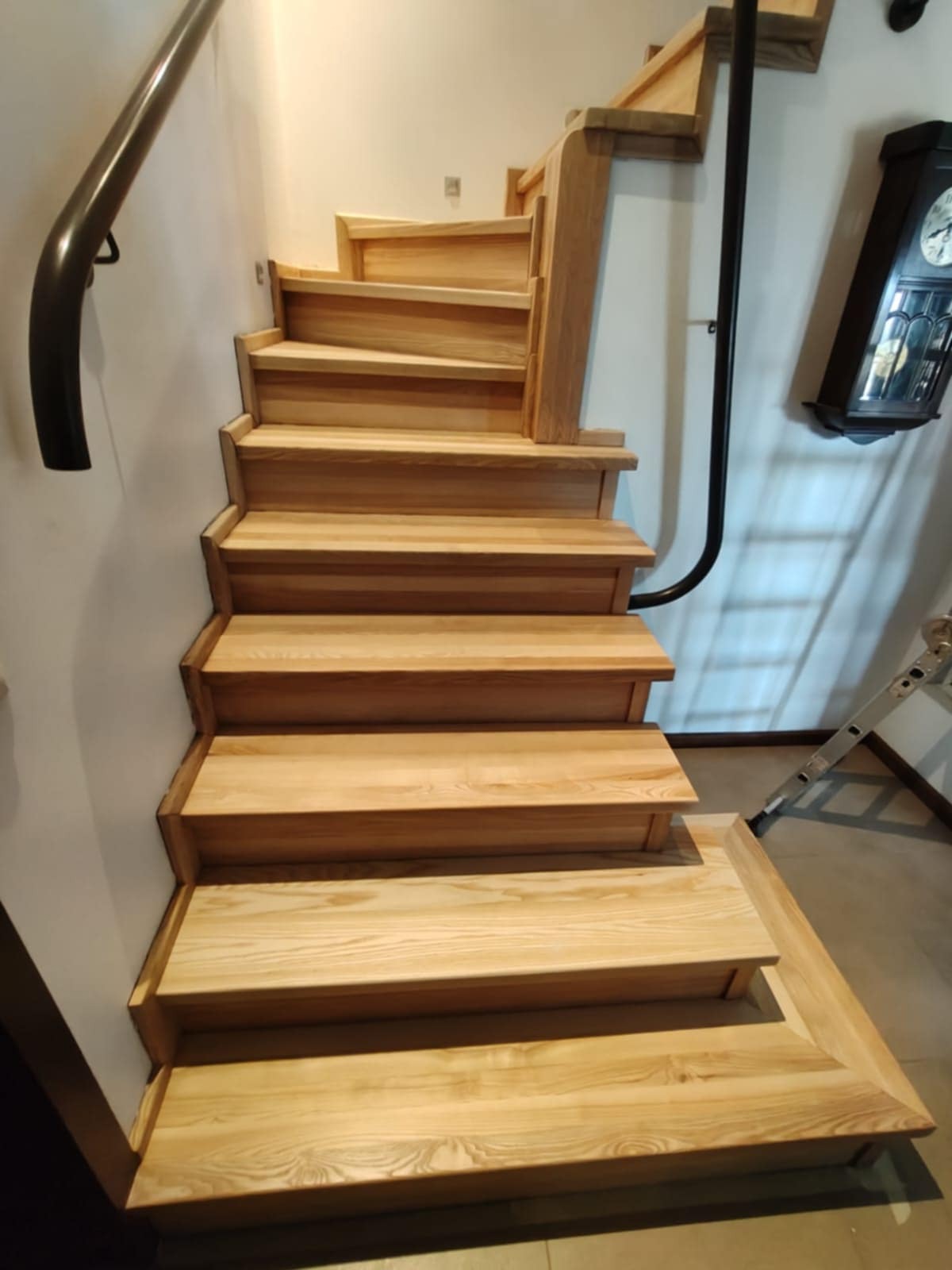 Jasne, drewniane schody z czarną, metalową poręczą, prowadzące w górę. Widoczny fragment ściany i zegar na ścianie. Solidne wykonanie, naturalne wykończenie drewna.