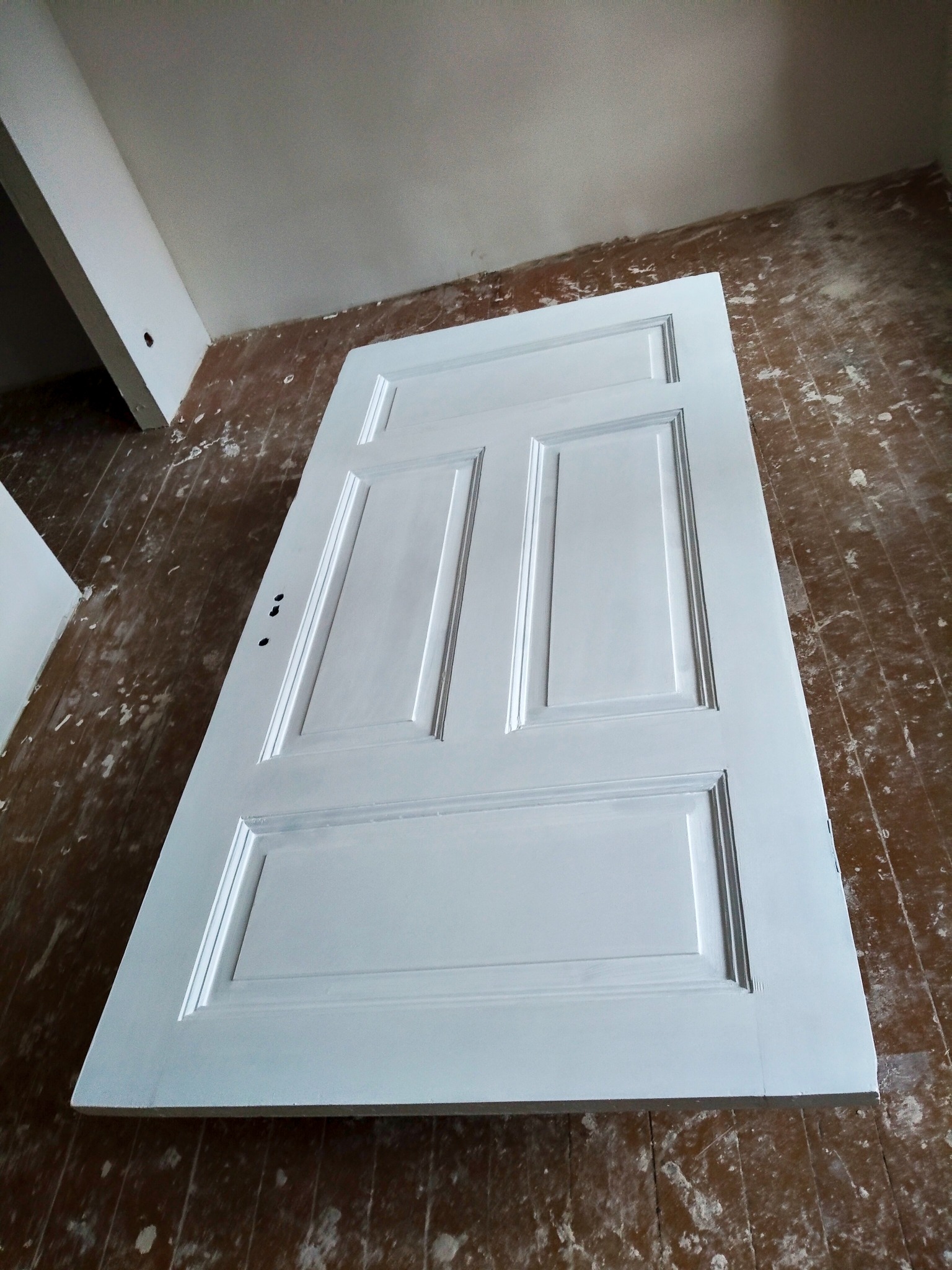 Białe, odnowione drzwi z płycinami leżące na podłodze z widocznymi śladami renowacji. Widoczne trzy otwory na klamkę. Ujęcie z góry, ukośne.