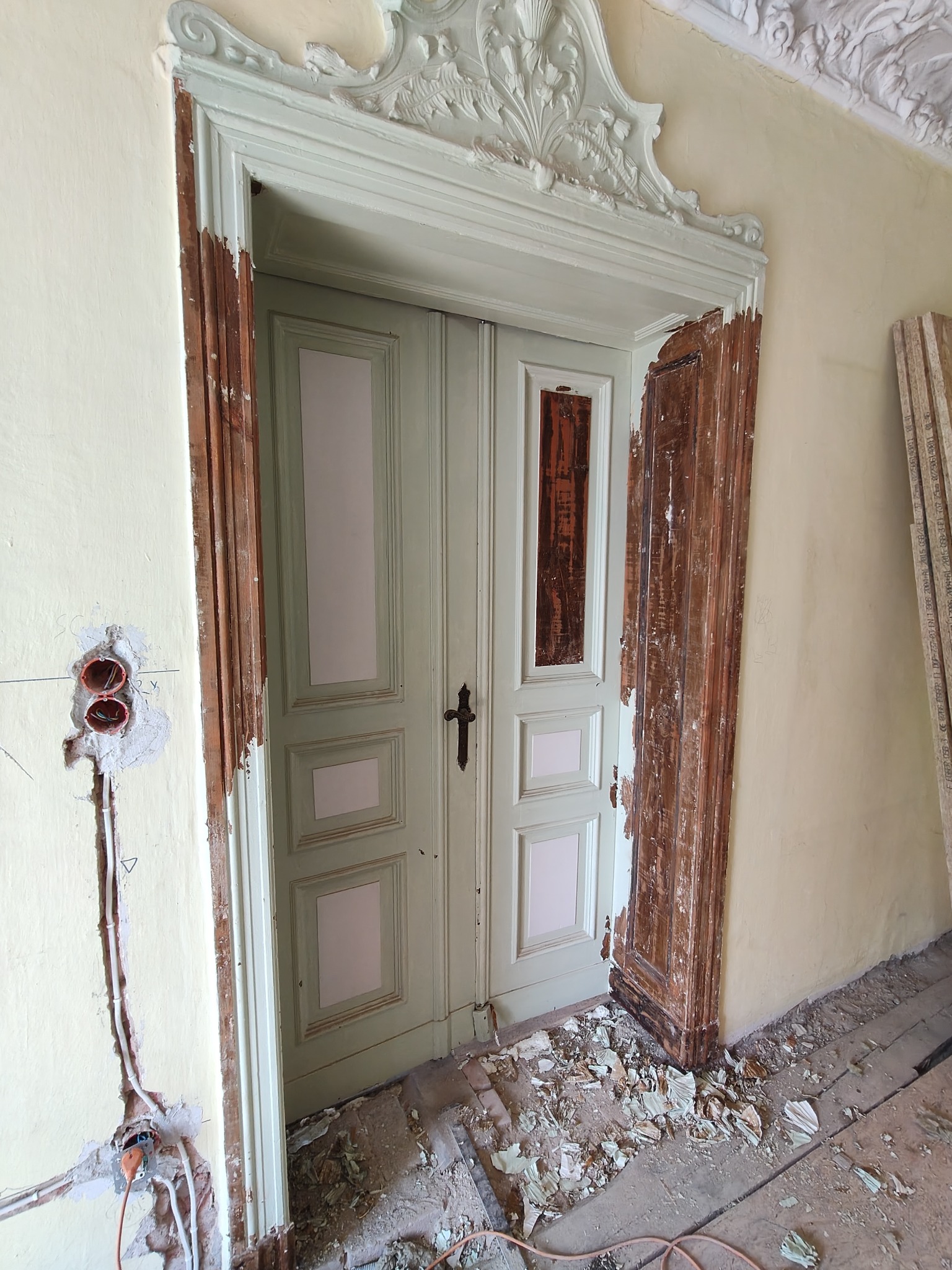 Renowacja zabytkowych drzwi z ozdobną ramą. Widoczne ślady po usuniętej farbie, odsłonięte drewno i przygotowane podłoże pod instalację elektryczną.