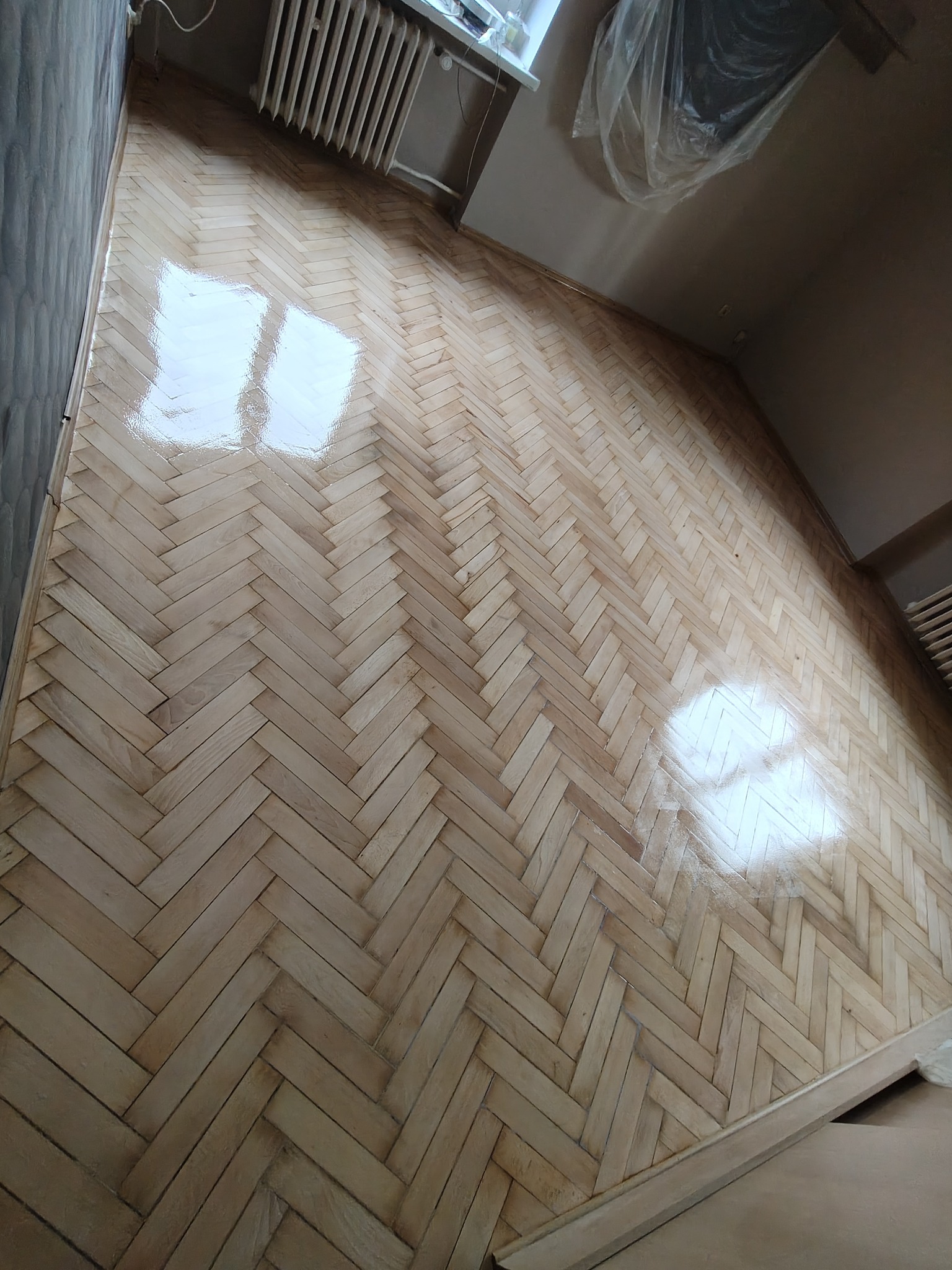 Parkiet ułożony w jodełkę po renowacji, z widocznym połyskiem lakieru. Grzejnik i fragment ściany w tle. Widoczne odbicia światła na powierzchni podłogi.