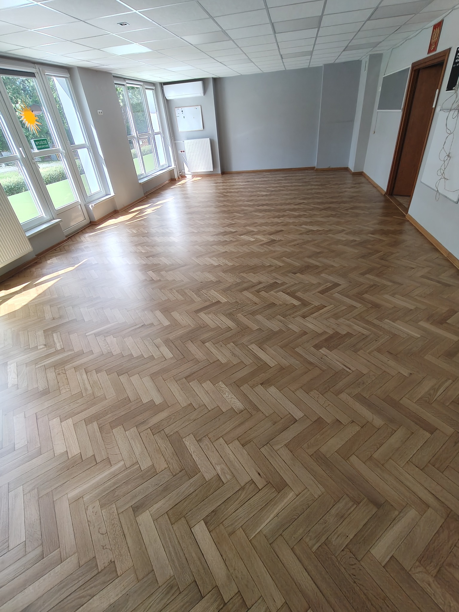 Jasny parkiet ułożony w jodełkę w przestronnym, słonecznym pomieszczeniu z oknami. Widoczne ślady po cyklinowaniu, podkreślające naturalny rysunek drewna.