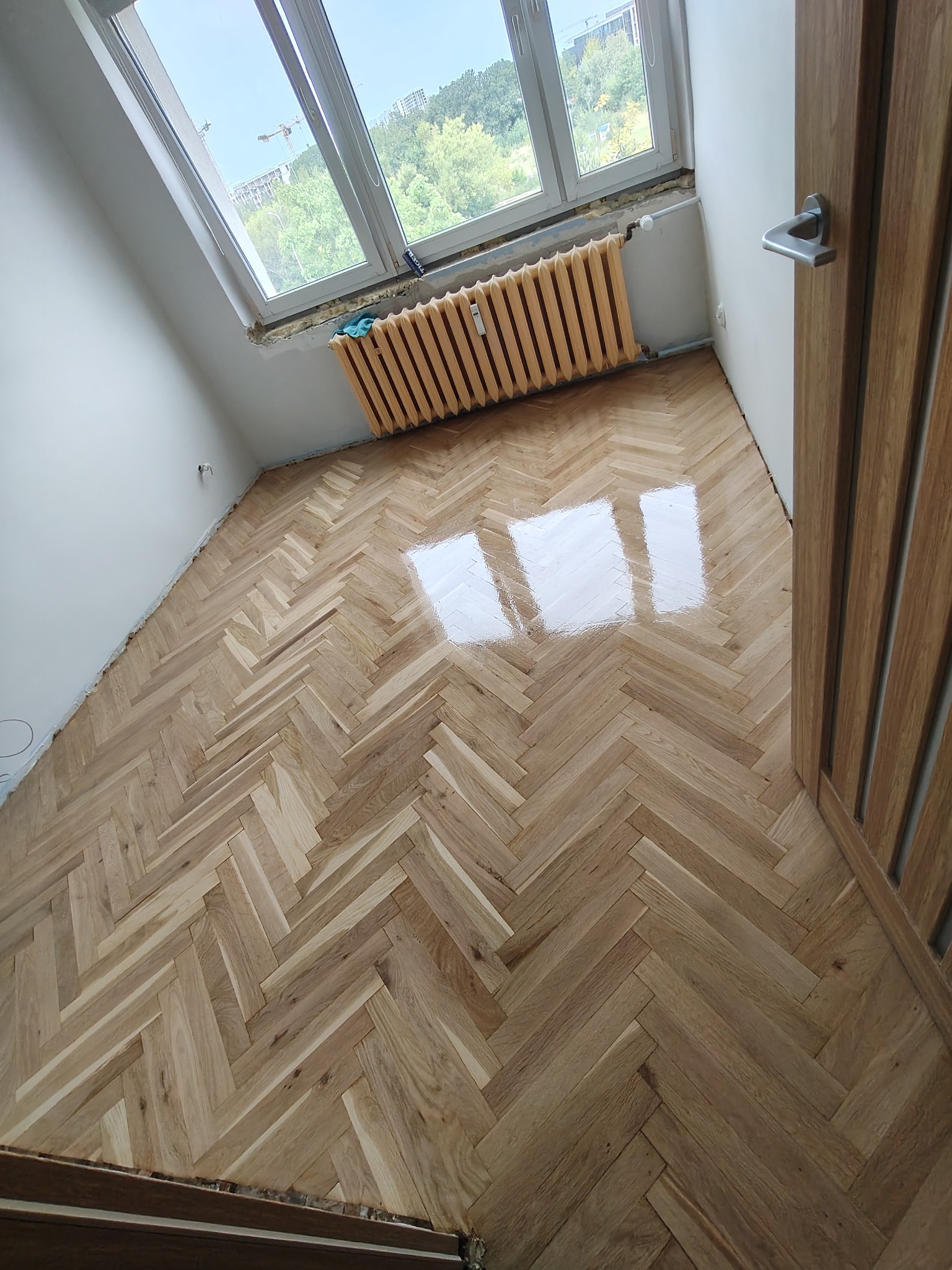 Parkiet ułożony w jodełkę, świeżo polakierowany, z widocznymi refleksami światła. Grzejnik pod oknem i drewniane drzwi w tle. Widok z góry.