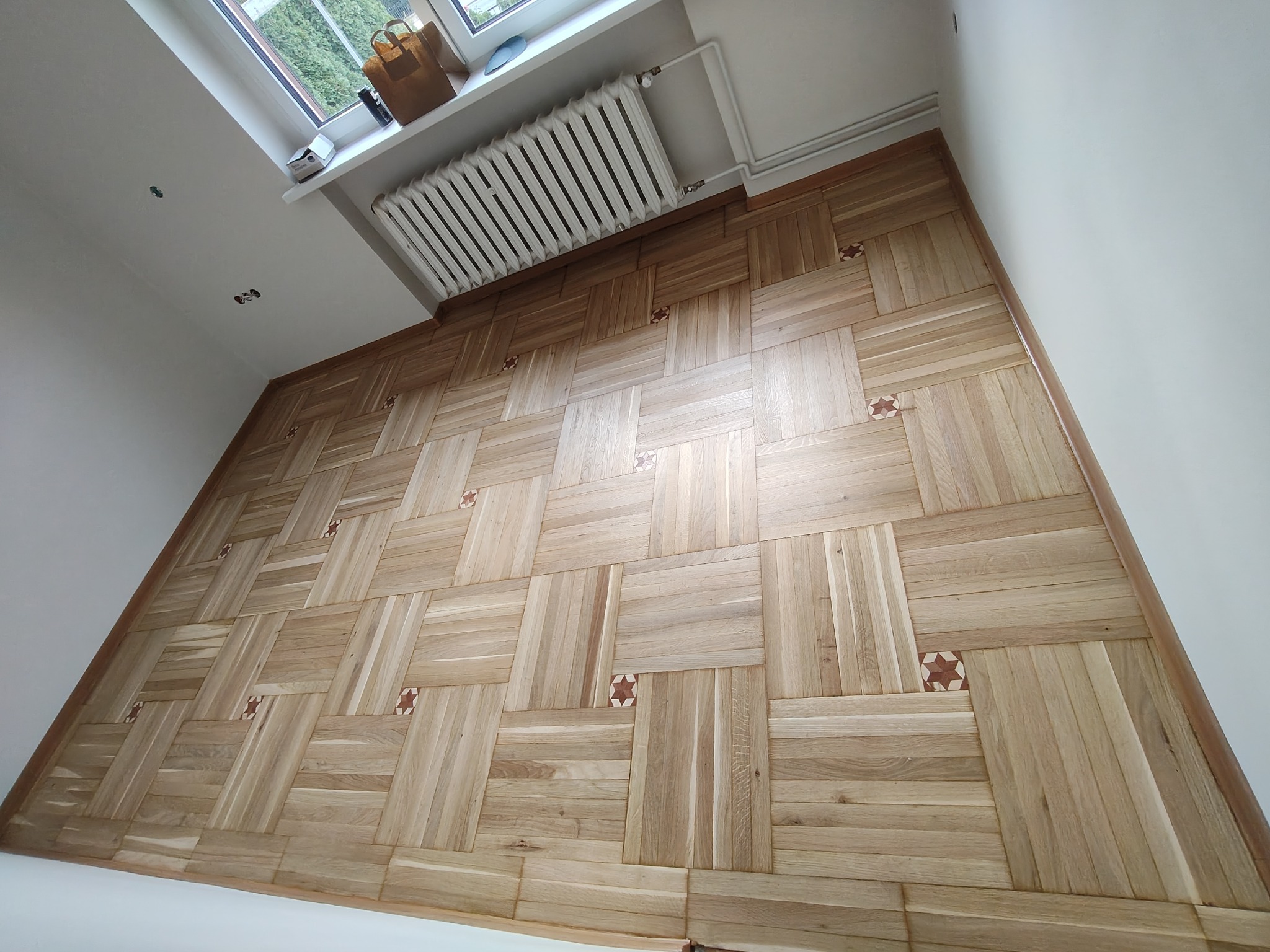 Parkiet z jasnego drewna ułożony we wzór koszykowy z dekoracyjnymi wstawkami w kształcie gwiazd, widok z góry, fragment grzejnika i okna w tle.