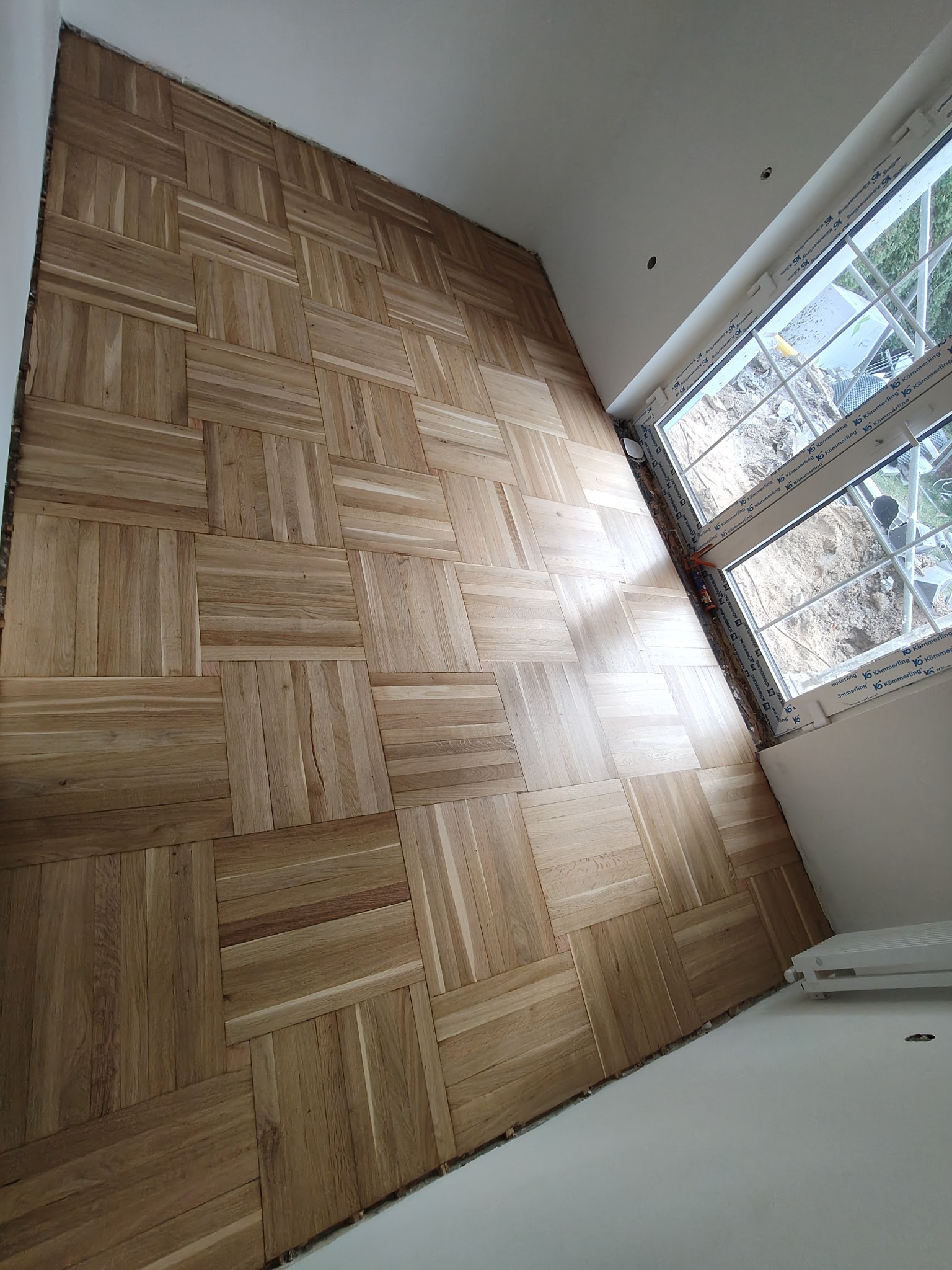 Parkiet ułożony w kwadraty, widok z góry, jasne drewno, świeżo po cyklinowaniu, widoczne okno z białymi ramami, ściany w trakcie wykańczania.