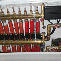 Daniel Tworzydlo - Rozdzielnia ogrzewania podłogowego z automatyką i sterowaniem, widoczne rury w izolacji, zawory, przepływomierze i moduł sterujący.