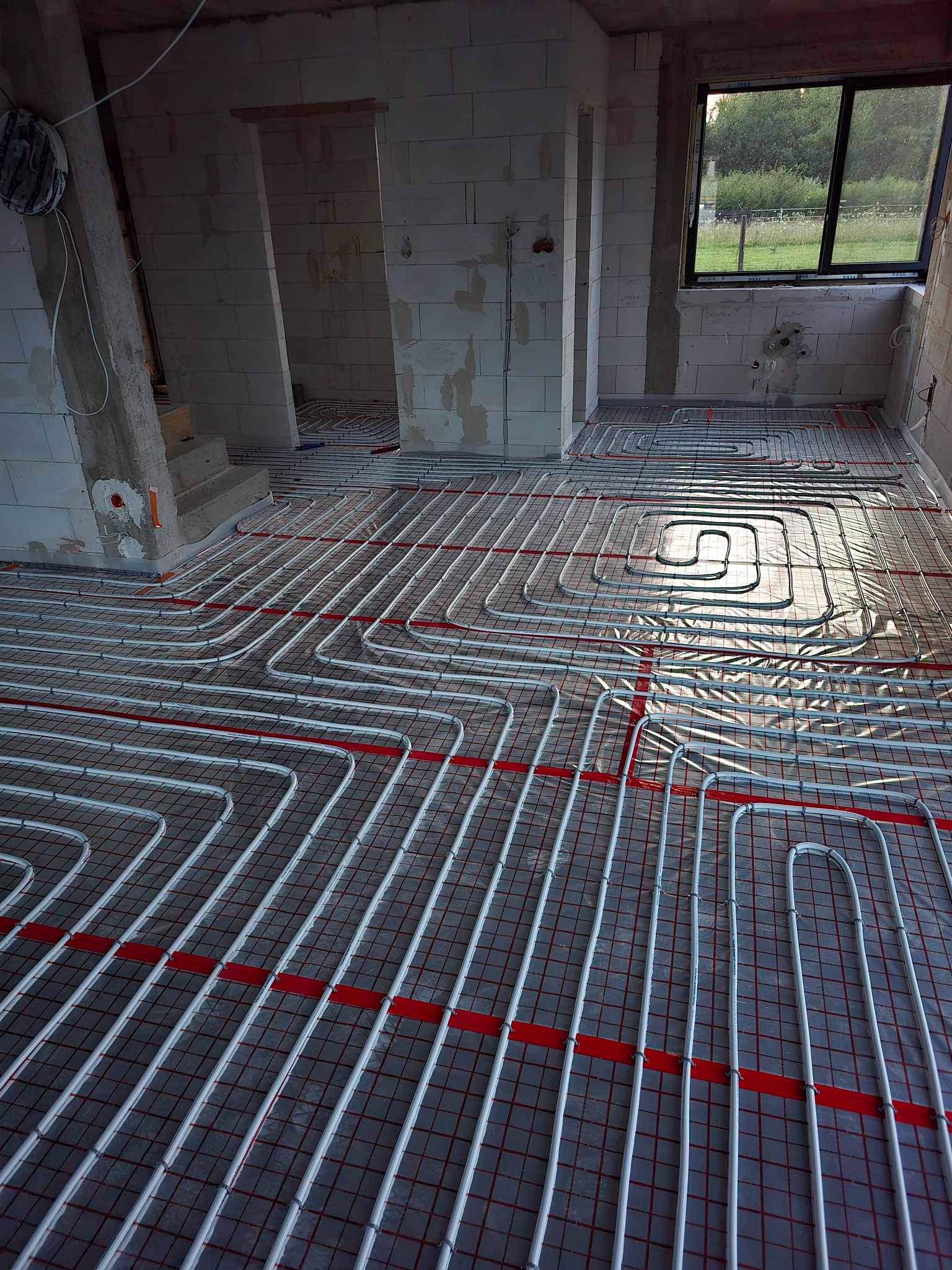 Białe rury ogrzewania podłogowego ułożone na czerwonej siatce w surowym wnętrzu. Widoczne okno i ściany z betonu komórkowego.