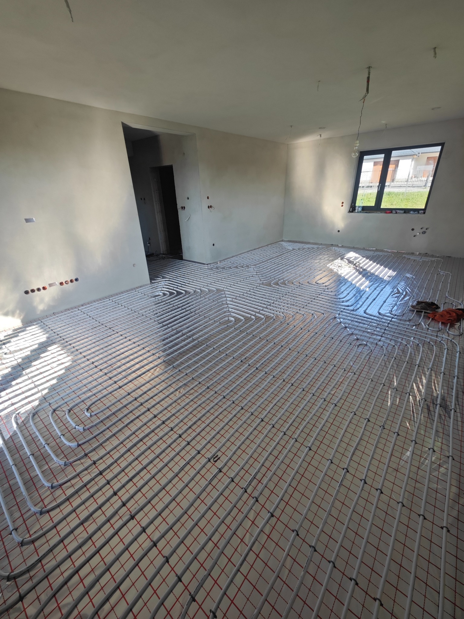Białe rury ogrzewania podłogowego na czerwonej siatce w surowym wnętrzu z oknem i nieotynkowanymi ścianami. Widoczne narzędzia i buty robocze.