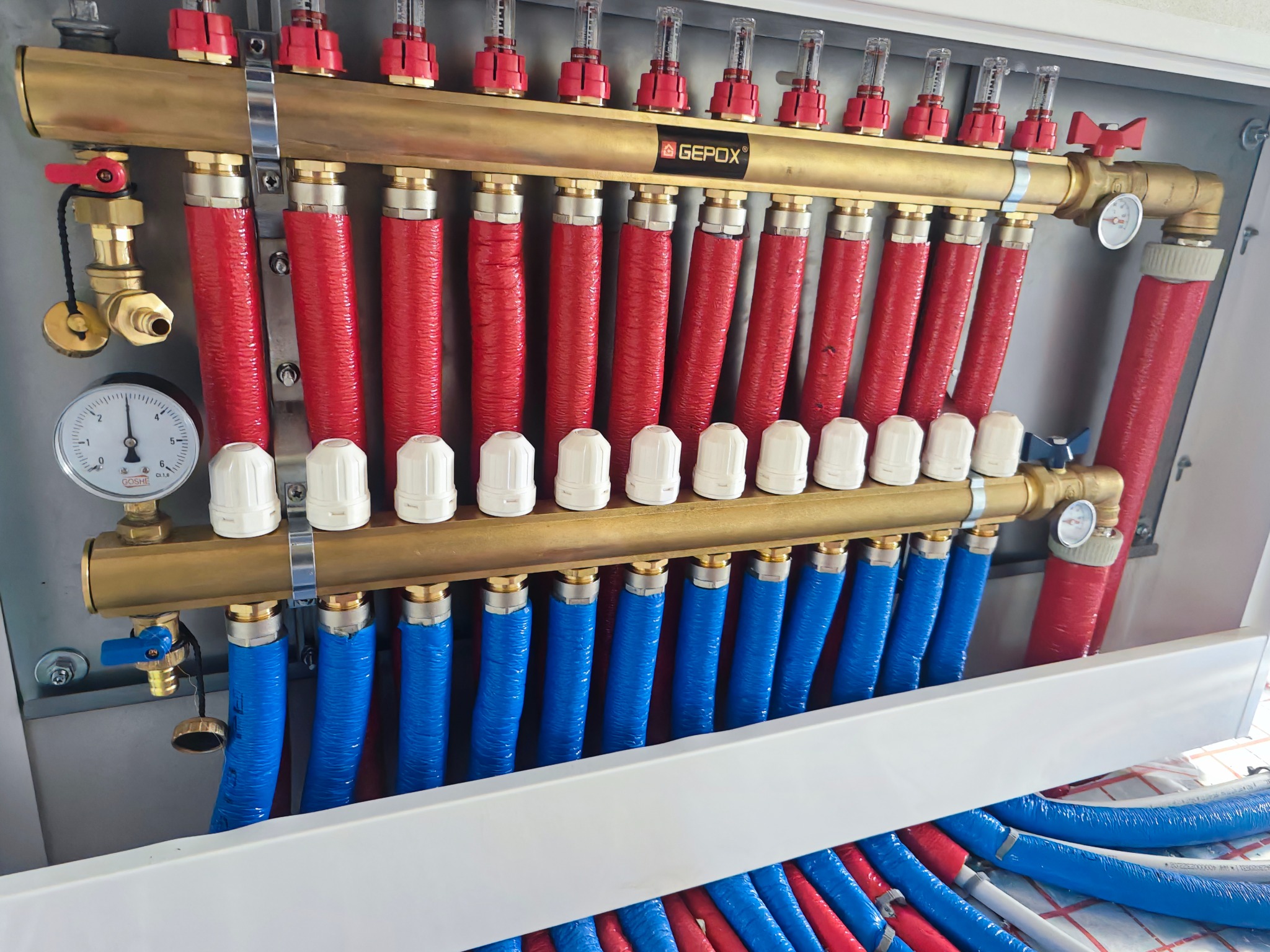 Rozdzielacz ogrzewania podłogowego GEPEX z precyzyjnymi przepływomierzami, czerwonymi i niebieskimi rurami. Widoczny manometr i zawory. Całość w szarej obudowie.