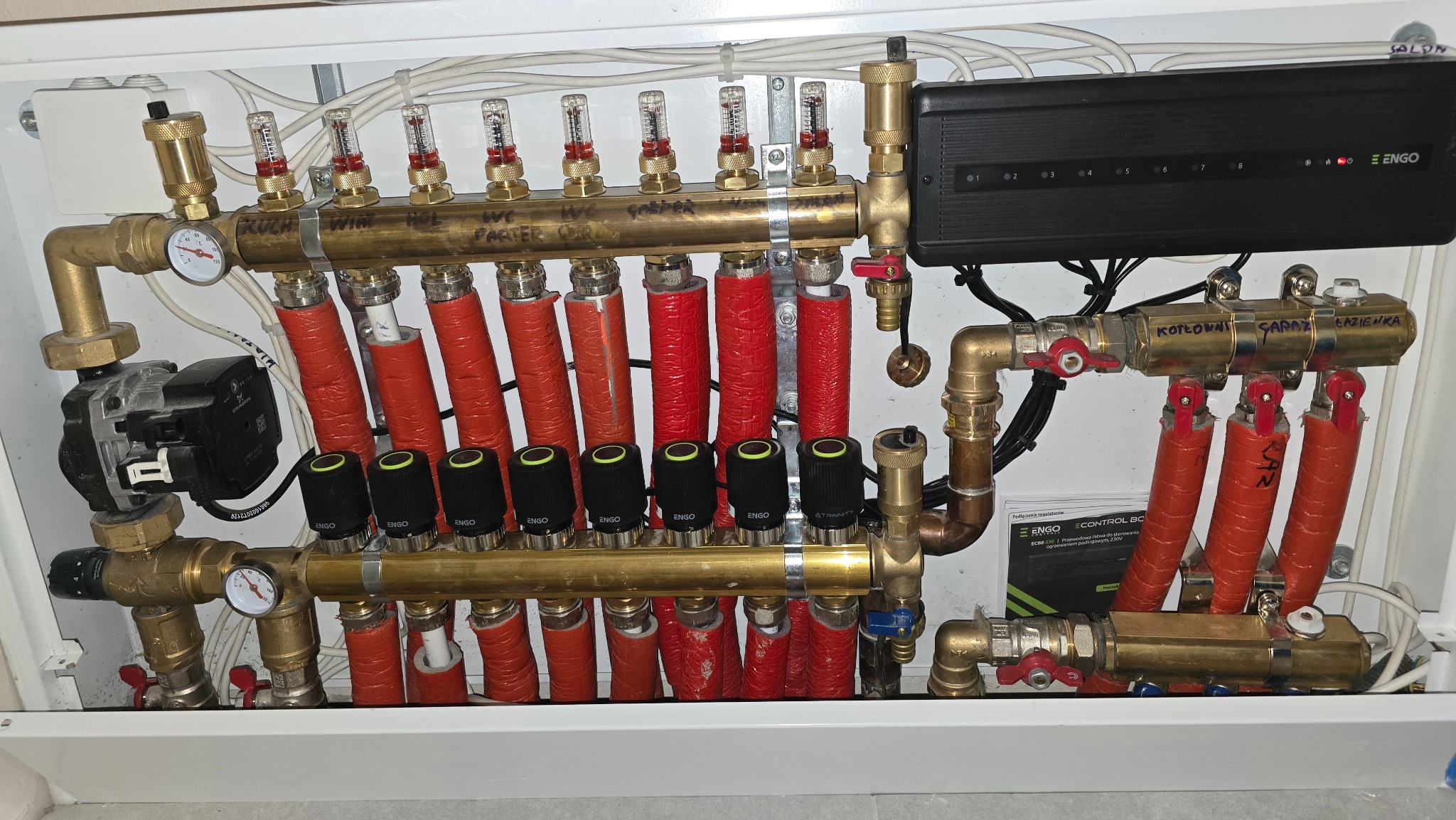 Rozdzielnia ogrzewania podłogowego z automatyką i sterowaniem, widoczne rury w izolacji, zawory, przepływomierze i moduł sterujący.
