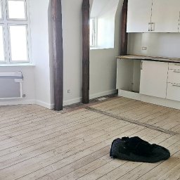 MADO Mateusz Dowejko - Wnętrze w trakcie remontu: białe ściany, drewniane belki, jasna podłoga z desek, fragment białej zabudowy kuchennej. Na podłodze leży czarna kurtka.