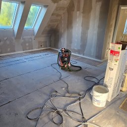 MADO Mateusz Dowejko - Pokój na poddaszu w trakcie szpachlowania ścian i sufitu. Widoczne okna dachowe, narzędzia pracy: wiadro z farbą, szpachelka, odkurzacz przemysłowy i kable.