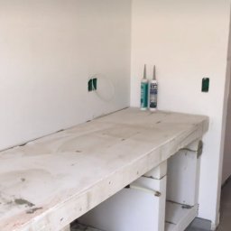 MADO Mateusz Dowejko - Wstępny etap montażu blatu kuchennego z betonu architektonicznego na białych wspornikach. Widoczne tuby silikonu i instalacje elektryczne w ścianie.