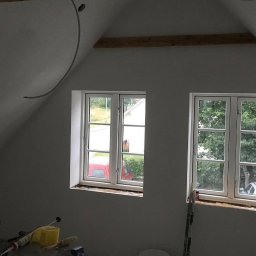 MADO Mateusz Dowejko - Poddasze po szpachlowaniu, przygotowane do malowania. Dwa okna z widokiem na zieleń. Widoczne narzędzia malarskie i listwa oświetleniowa na suficie.