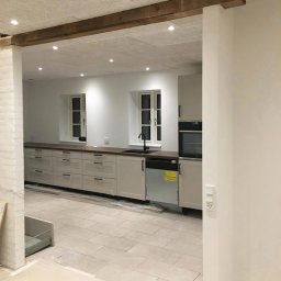 MADO Mateusz Dowejko - Jasna kuchnia w trakcie wykończenia: kremowe szafki z ciemnym blatem, białe ściany z oknami i drewniana belka stropowa. Widoczny fragment podłogi z płytek.