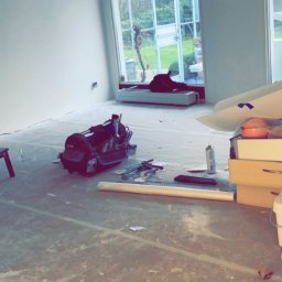 MADO Mateusz Dowejko - Wnętrze pokoju w trakcie prac wykończeniowych: widoczne narzędzia, torba narzędziowa, folia ochronna na podłodze, ściany po szpachlowaniu, duże okna z widokiem na zieleń.