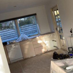 MADO Mateusz Dowejko - Remont kuchni w trakcie: zabezpieczone folią meble, okno i podłoga. Widoczne narzędzia malarskie i puszka z farbą. Ściany przygotowane do malowania.