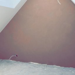 MADO Mateusz Dowejko - Poddasze ze ścianą pokrytą świeżą, brązową gładzią. Widoczne okno dachowe z drewnianą belką i wystające kable elektryczne. Podłoga przygotowana do dalszych prac wykończeniowych.