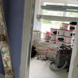 MADO Mateusz Dowejko - Wnętrze w trakcie prac wykończeniowych: ściana z płyt gipsowych, drabina malarska, wiadro z farbą, odkurzacz przemysłowy. Widoczne elementy konstrukcji sufitu.