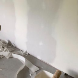 MADO Mateusz Dowejko - Ściana po szpachlowaniu, gotowa do malowania. Widoczne narzędzia: wałek w wiadrze z farbą i taśmy malarskie na podłodze. Przygotowanie do malowania ścian.