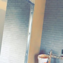 MADO Mateusz Dowejko - Montaż metalowej ościeżnicy w surowym wnętrzu. Widoczna ściana z nałożoną warstwą gładzi, drabina malarska i wiadro z zaprawą. Ujęcie z dołu, podkreślające wysokość pomieszczenia.