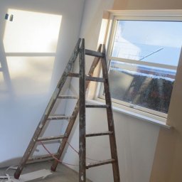 MADO Mateusz Dowejko - Pokój na poddaszu w trakcie malowania: drewniana drabina oparta o ścianę, częściowo pomalowana na beżowo, okno w tle, resztki taśmy malarskiej na podłodze.