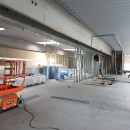 MADO Mateusz Dowejko - Wnętrze w trakcie wykończenia: metalowe profile ścian działowych, izolacja w rolkach, podnośnik nożycowy. Widoczny fragment sufitu z instalacjami.